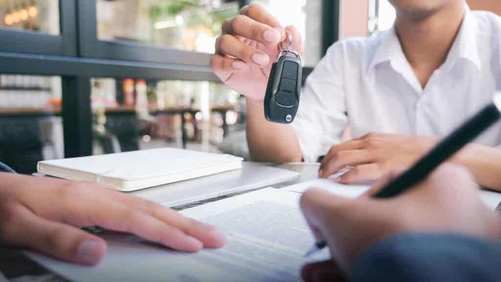 woman handing car keys to buyer as he signs loan paperwork