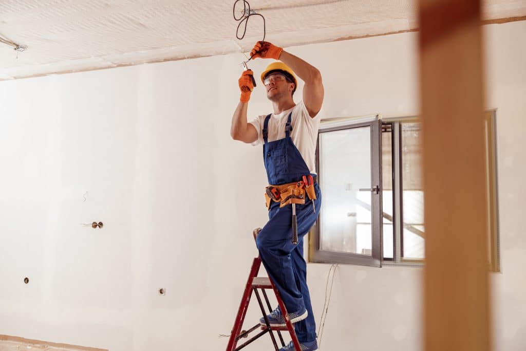 electrician in overalls on ladder fixing wires in the ceiling