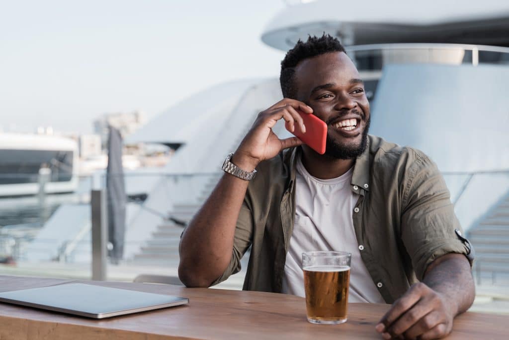 Young african man drinking beer at brewery bar using mobile phone with luxury yacht port in background