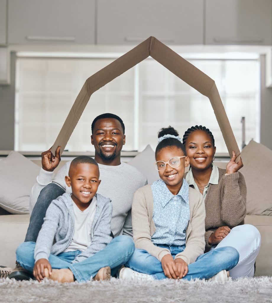 family sitting under piece of cardboard shaped like a house